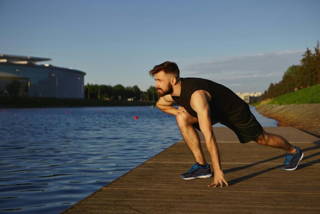 A man is doing squats on a dock by the water.
